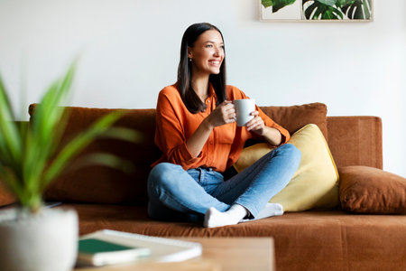 Happy European lady sitting on comfortable sofa, drinking coffee and looking away, thinking about plans, resting in modern cozy living roomの写真素材