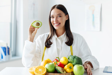 Cheerful nutritionist with slice of avocado smiling at camera, sitting at office with wooden board full of fruits and vegetablesの写真素材