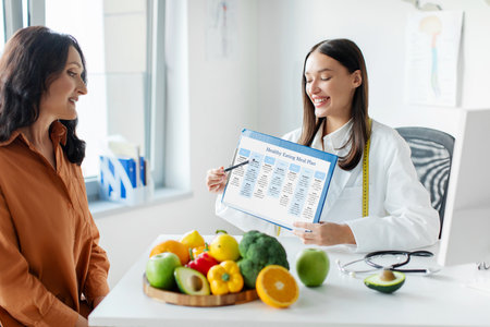 Positive young woman dietologist showing mature female patient medical chart, discussing weekly menu plan, having consultation in officeの写真素材