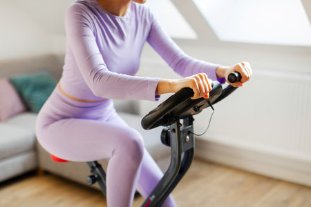 Sports woman in sportswear cycling bike at home in light living room, cropped shot. Domestic cardio...