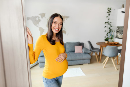 Beautiful pregnant woman touching her belly and smiling while standing near opened door and inviting to her apartmentの写真素材