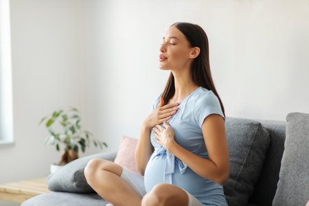 Pregnant woman doing yoga breathing exercises with eyes closed, sitting on couch at home. Relaxation and childbirth healthcare conceptの写真素材