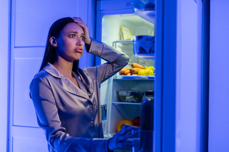 Confused young woman in pajamas looking in open refrigerator full of products and touching head, standing near fridge late at nightの写真素材