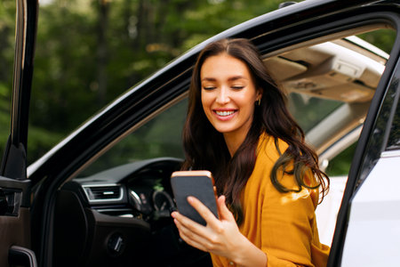 Positive woman getting out of her car and using cellphone, sitting with opened door, stopping for...