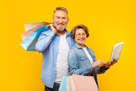 Cheerful elderly man and woman shopping on Internet together, using laptop and holding purchases, posing over yellow backgroundの写真素材