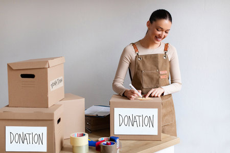 Donation concept. Woman volunteer writing address on sticker and sticking it on donate box with full...