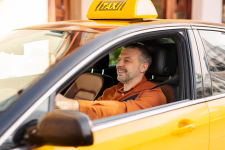 Photo of middle aged driver man sitting in yellow taxi, driving on city street with smile, holding hands on steering wheelの写真素材