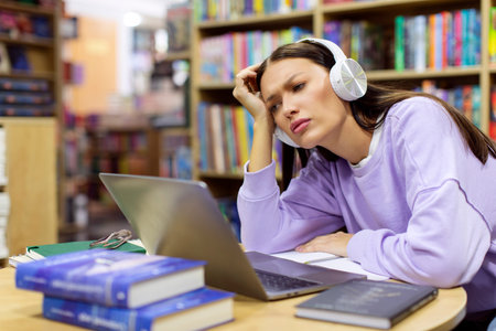 Fatigued tired female student overworking on study project in library, sitting at table with laptop...