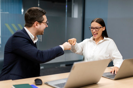 Happy employees fist bumping in celebration of sales goals or successful teamwork results, sitting at office desk. Support, partnership growth, achievement conceptの写真素材