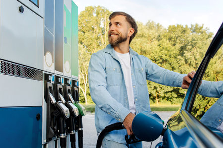 Best fuel price. Positive man refueling automobile tank with bio fuel, standing with gun nozzle near petrol dispenserの写真素材
