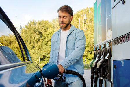 Bearded man refilling his auto with fuel at petrol station, holding filling gun nozzle. Automobile...
