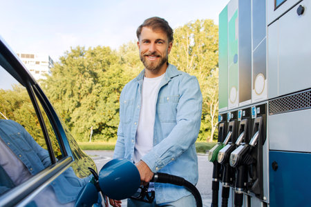 Eco friendly refueling concept. Handsome bearded man filling up his automobile at modern gas or petrol station, smiling at cameraの写真素材
