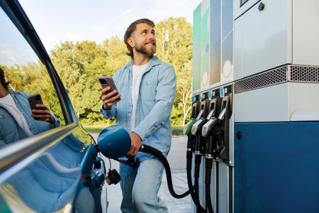 Bearded man refueling car and using smartphone, comparing fuel prices on petrol meter screen and gas...