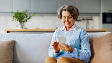 Happy elderly woman in glasses reading text message on cellphone, browsing Internet, using online...