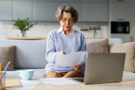 Focused senior woman sitting at table, using laptop, banking online or checking utility bills, financial statement papers, received letter from bankの写真素材