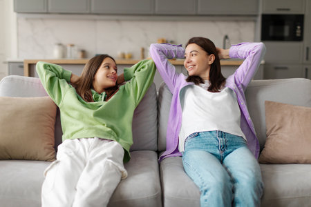 Mother and teen daughter chilling at home on sofa, relaxing with hands behind head and smiling at each other, having pleasant conversationの写真素材