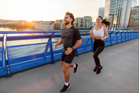 Young athlete couple doing workout outdoors, man and woman running on footbridge in the evening with sunset on backgroundの写真素材