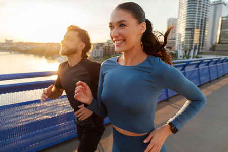 Sporty European couple in sportswear running together while working out outdoors on city bridge