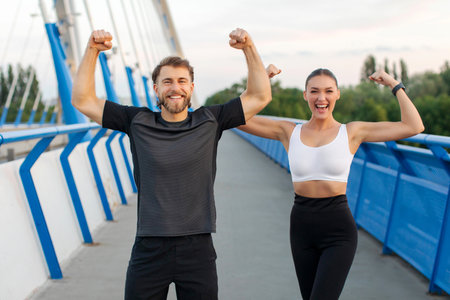 We did it. Excited sporty couple doing sport together outdoors, showing biceps and rejoicing sport achievement, standing on city footbridgeの写真素材