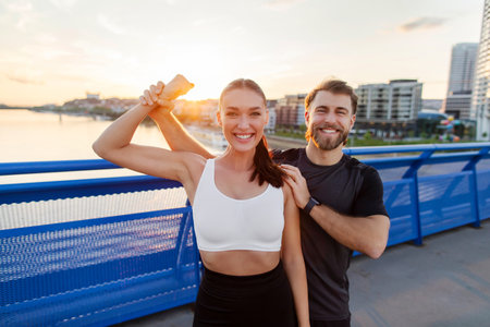 Happy sporty woman showing biceps, posing with husband after workout outdoors on city bridge, having running training in the eveningの写真素材