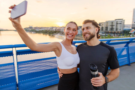 Romantic European couple making selfie on smartphone after evening run and outdoors workout, standing on bridgeの写真素材