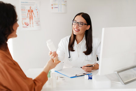 Drug prescribing. Positive doctor woman giving jar of pills to patient and giving instructions on medication treatment, sitting in office during consultationの写真素材