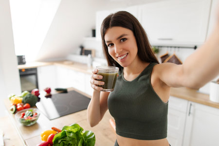 Happy lady making selfie posing with smoothie after morning workout, standing in kitchen interior at home, enjoying green detox drink caring for healthの写真素材