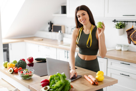 Fitness nutritionist lady at table with laptop computer, holding apple, posing with measure tape in kitchen at home, recommending healthy nutrition, copy spaceの写真素材