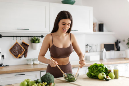 Happy young sportswoman cooking fresh green salad in modern kitchen, mixing ingredients in bowl, enjoying balanced dietの写真素材