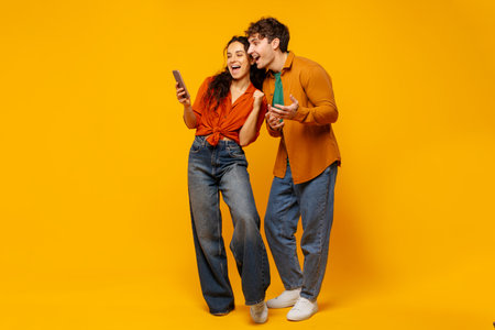 Overjoyed couple looking at smartphone with joy and excitement, reading great news or celebrating online win over yellow studio backgroundの写真素材