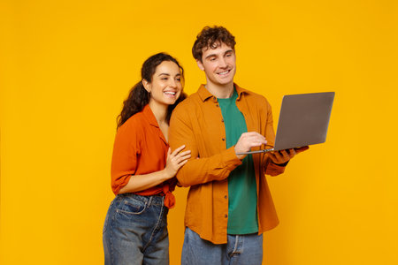 Cheerful European couple using modern pc laptop and smiling at camera, posing together on orange studio backgroundの写真素材