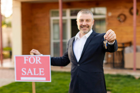 Real estate agent selling house and real estate, man giving new house key, standing near for sale sign and smiling at cameraの写真素材
