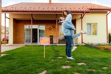 Property and purchase concept. Happy spouses new homeowners posing in front of their house with sold sign nearby, full length shotの写真素材