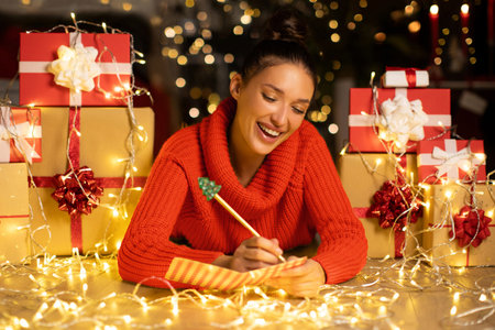 Excited European lady writing greeting card or letter to Santa, lying on floor among gift boxes and garland lightsの写真素材