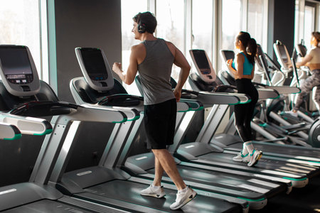 Strong man running on treadmill in well-lit gym, wearing workout clothes and headphones, focused on...