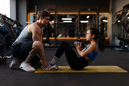 Male trainer assisting young lady with her workout in gym, woman practicing sit-ups while holding...