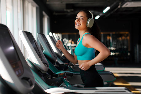 Positive sports woman working out and running on treadmill in modern gym, listening music in...