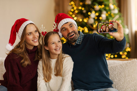 Cheerful family wearing Santa hats taking selfie with smartphone, sitting on couch in cozy living room, decorated with sparkling Christmas tree on backgroundの写真素材