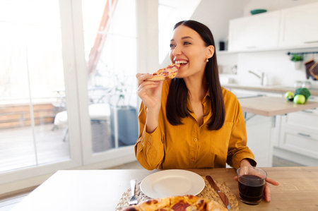 Happy lady enjoying tasty pizza, holding and biting slice, sitting at table in kitchen interior, copy spaceの写真素材
