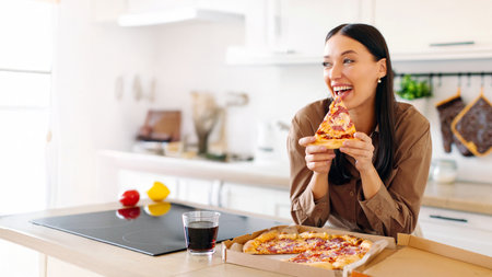 Excited lady holding slice of tasty pizza, enjoying her meal, standing at kitchen counter with carton box from delivery serviceの写真素材