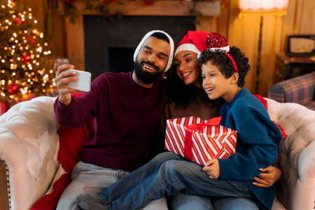 Happy African American family of three capturing selfie at Christmas Eve, sitting on couch,...