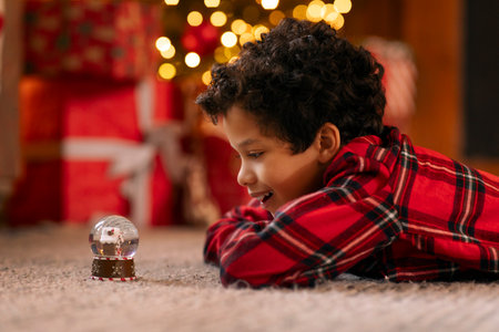 Happy Latin boy in red plaid shirt lying on the floor carpet, captivated by shimmering snow globe, side viewの写真素材