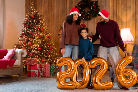 Happy New Year. Latin family with child standing near Christmas tree, wearing Santa hats and holding large golden balloons with 2026 numberの写真素材