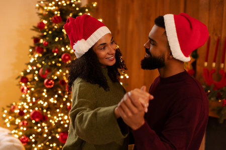Loving black couple in Santa hats dancing in cozy room with decorated Christmas tree, glowing lights and warm festive atmosphereの写真素材