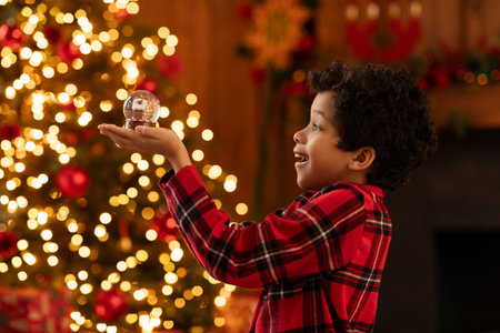 Overjoyed black boy in red checkered shirt smiling as holding snow globe, admiring its shimmering details, beautifully decorated Christmas tree glowing with warm lights on backgroundの写真素材