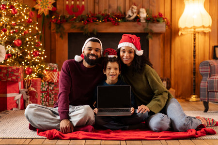Cheerful black family sitting on blanket in warmly decorated living room for Christmas, showing laptop blank screen, surrounded by gifts and holiday decorations, mockupの写真素材