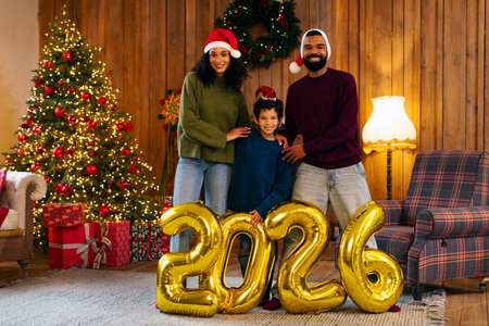 Latin family of three standing in front of Christmas tree with golden balloons marking 2026 New Year, posing in room filled with festive cheer, free spaceの写真素材