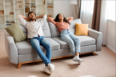 Calm Indian couple resting on couch with hands behind their heads, embodying serene and blissful domestic moment of togetherness and comfortの写真素材