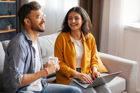 Couple sitting on sofa with laptop and coffee mug, woman using computer while man drinking tea, browsing internet or shopping online togetherの写真素材