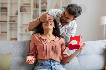 Indian guy closes eyes to surprised woman and giving present in box, lady sitting on couch in living room. Party at home, birthday gift, anniversary, Valentines day and holidayの写真素材